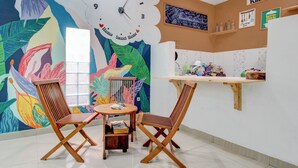 Een gezellig café met een kleurrijke muurschildering, een ronde houten tafel, twee houten stoelen, een klok en een keukentoog.