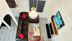 Una sala de estar con un sofá modular, una mesa de centro, una araña, un televisor y una ventana con persianas.