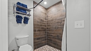 Un baño con un inodoro blanco, una ducha con piso de baldosas y un perchero con toallas azules.
