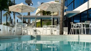 A swimming pool with white chairs and umbrellas, surrounded by modern buildings. - 119 Ambassador  (Joao Pessoa)