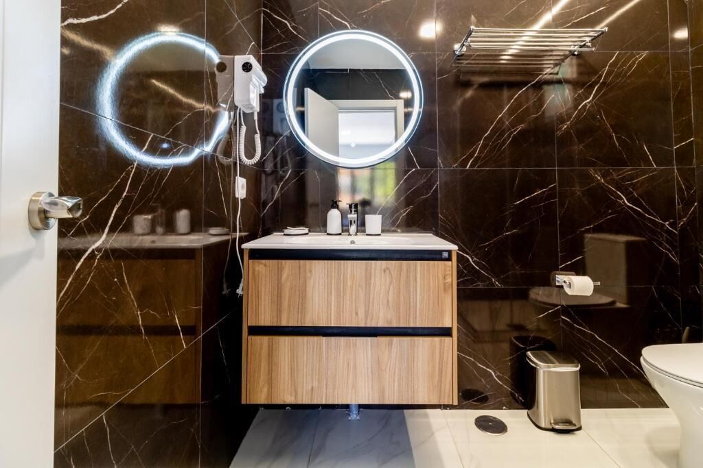 Een moderne badkamer met een marmeren wand, een houten wastafelmeubel, een ronde spiegel met verlichting en een toilet.