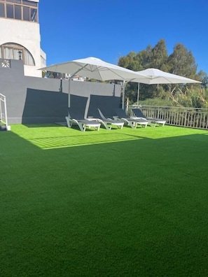 A pool area with artificial grass, lounge chairs, and umbrellas.