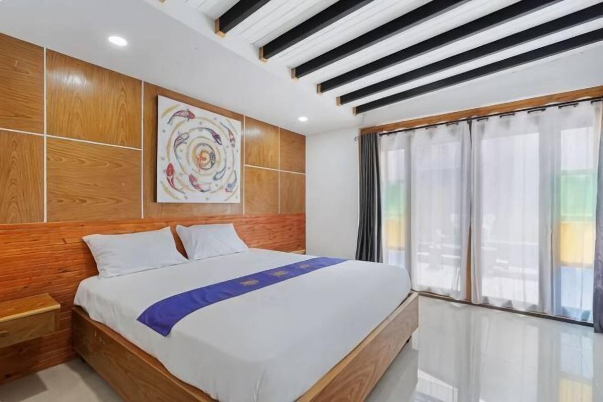 A hotel room with a wooden headboard, a bed with white linens, a blue bedspread, a wall-mounted artwork, and a large window with curtains.