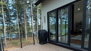 A wooden deck with a grill, a sliding glass door leading to an interior space, and a forested backdrop. - Modern Lakehouse on Quiet Peninsula (Lakeville)