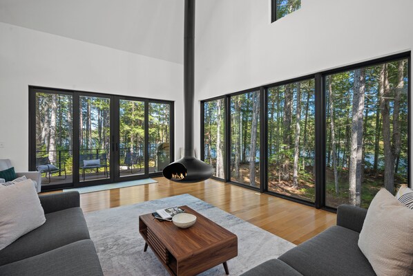 A spacious living room with large windows, a fireplace, and a view of a forest.