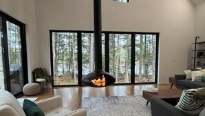 A modern living room with a fireplace, large windows, and a view of trees. - Modern Lakehouse on Quiet Peninsula (Lakeville)