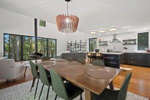 A modern kitchen with a central island, a dining area with a wooden table and green chairs, and large windows offering a view of trees. - Modern Lakehouse on Quiet Peninsula (Lakeville)