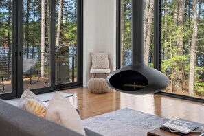 A modern living room with a fireplace, large windows, and a view of a forest.
