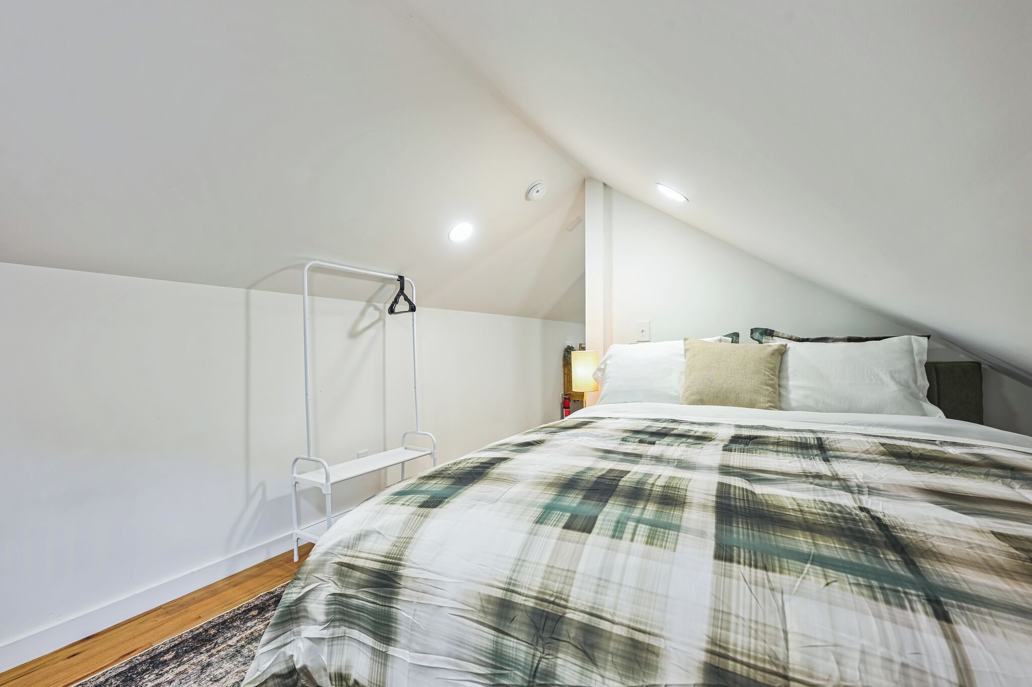 Un dormitorio con techo inclinado, una cama con colcha escocesa, una mesita de noche con lámpara y un radiador blanco.