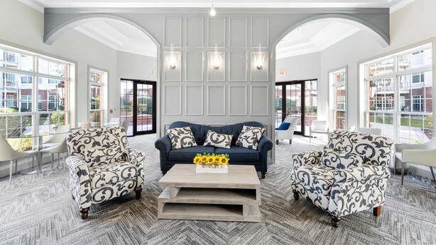 A spacious lobby with a dark blue sofa, patterned armchairs, and a coffee table with sunflowers. - Landing Apartments New York  (Raritan)