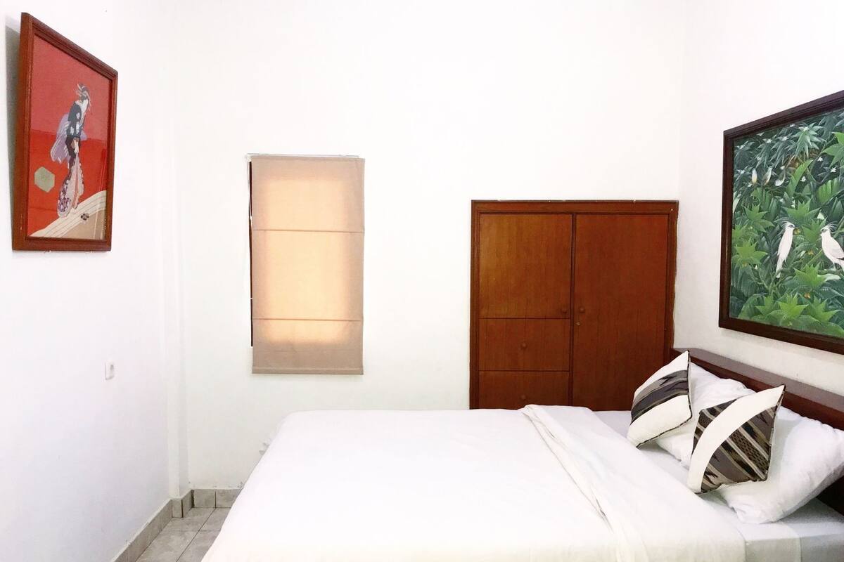 A bedroom with a bed, a wardrobe, a window with blinds, and two framed pictures on the wall.