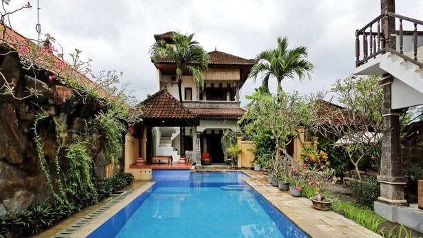 A swimming pool with a tiled edge, a house with a tiled roof, and a stone wall. - Rumah Akira Seminyak (Bali)