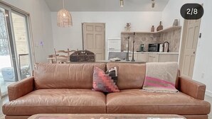 A living room with a brown leather sofa, a wooden coffee table with books and a plate of apples, and a kitchen in the background. - The Cork Cottages, Unit 1, Arcade Table, Modern and New 2b/2b (Geneva)