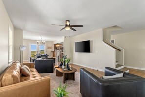 A spacious living room with a large sectional sofa, a dark leather couch, and a dining area with a wooden table and chairs. A ceiling fan and a flat-screen TV are mounted on the wall. - Large Home w wraparound porch/Amazing View/Hot Tub/Fire Pit (Morganton)