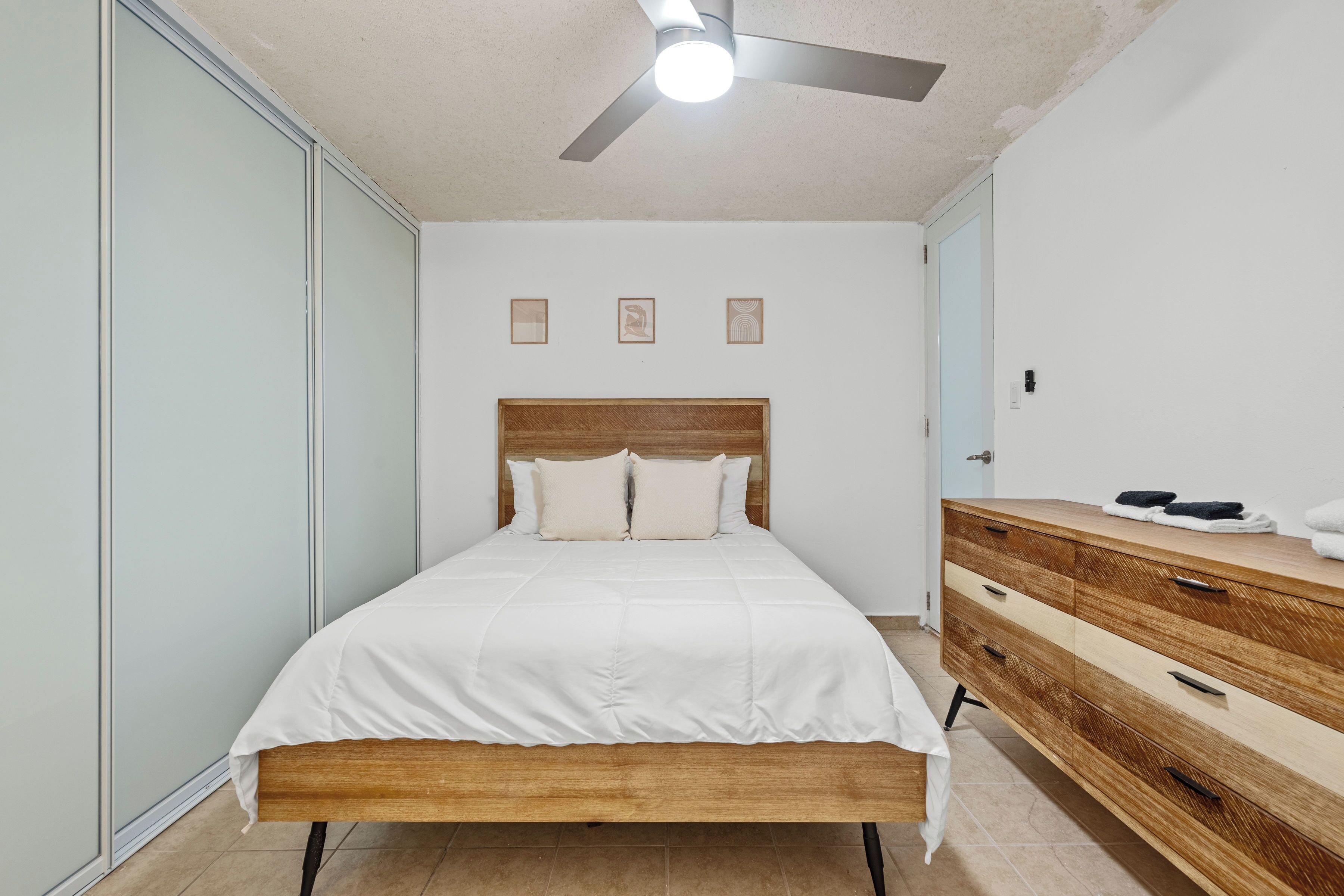 A bedroom with a bed, a wooden dresser, a ceiling fan, and a light fixture.