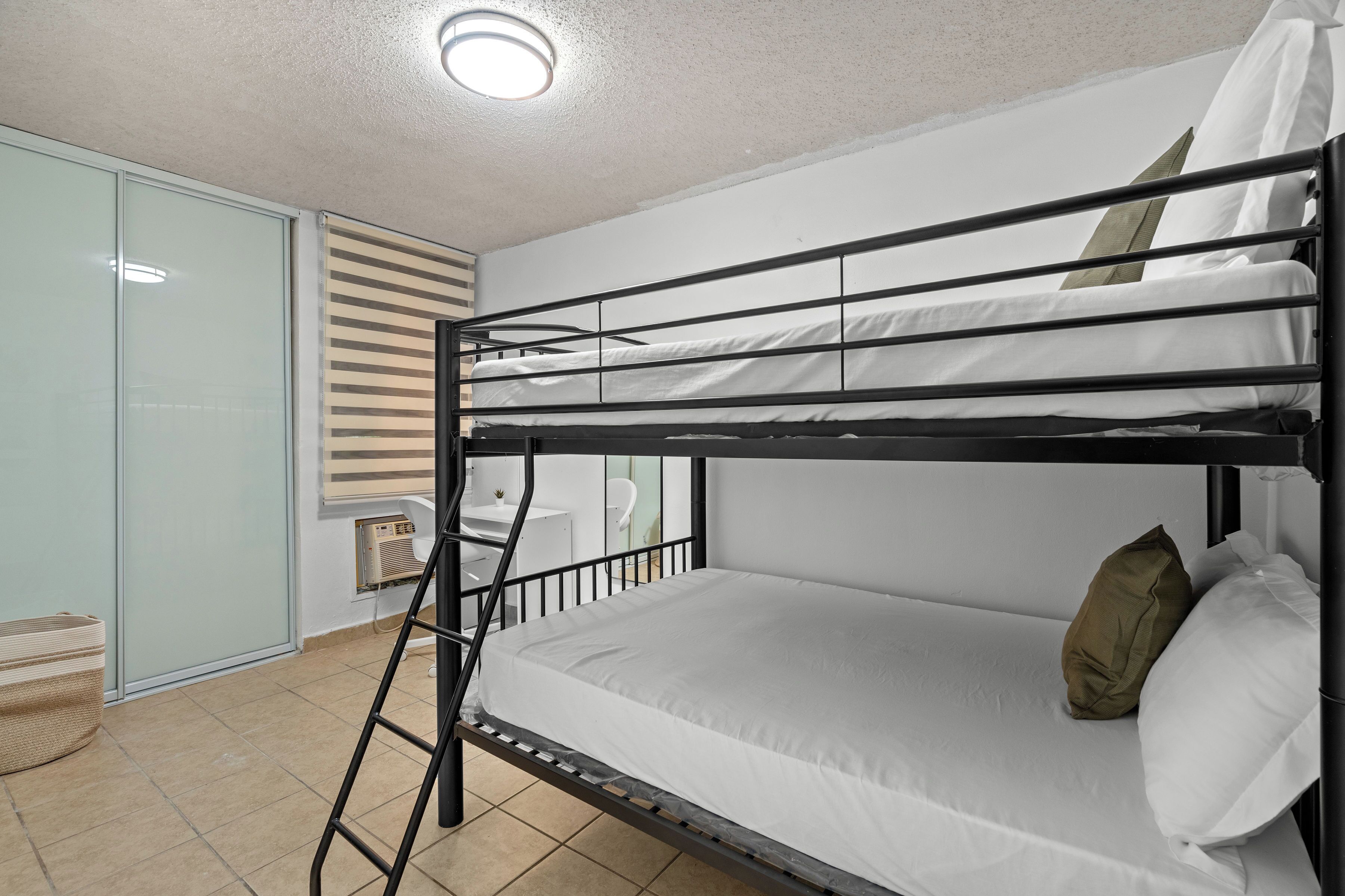 Un lit superposé avec une échelle, un matelas blanc et des oreillers. Il y a une petite table et une chaise dans le coin.