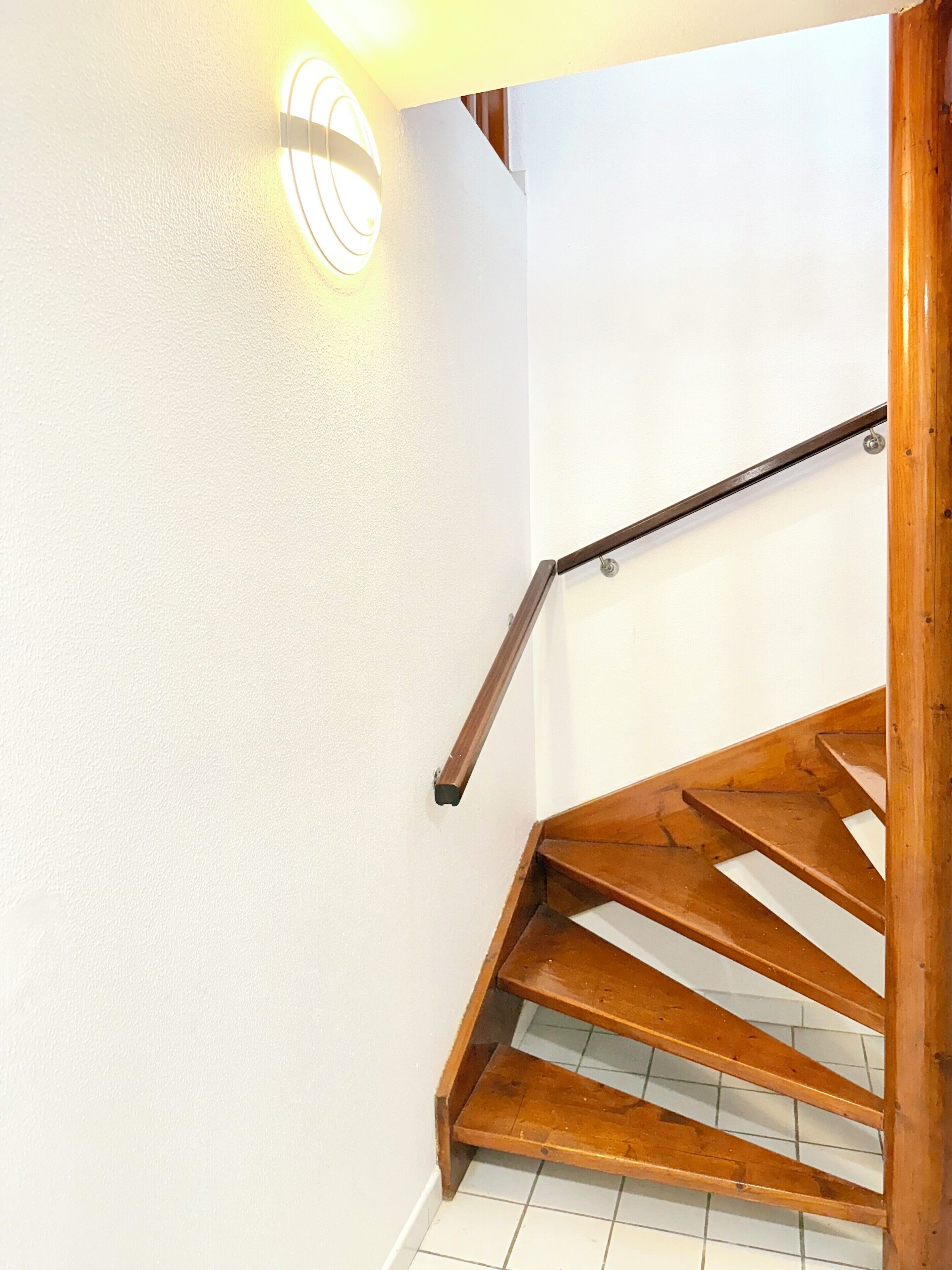 A wooden staircase with a handrail, white walls, and a light fixture on the wall.