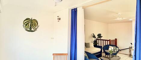 Una habitación con una mesa redonda, sillas, un ventilador de techo y vistas a otra habitación con una cama y un sillón.