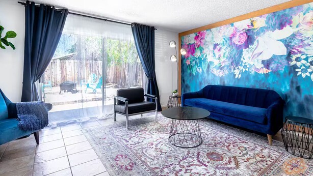 Ein Wohnzimmer mit einem blauen Sofa, einem schwarzen Sessel, einem runden Couchtisch und einem farbenfrohen Blumenfries an der Wand.