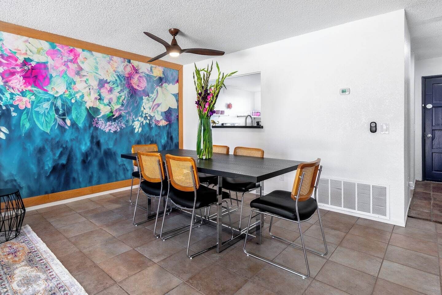 Un coin repas avec une table noire et quatre chaises, un ventilateur de plafond, un tableau floral aux couleurs vives, et une cuisine en arrière-plan.