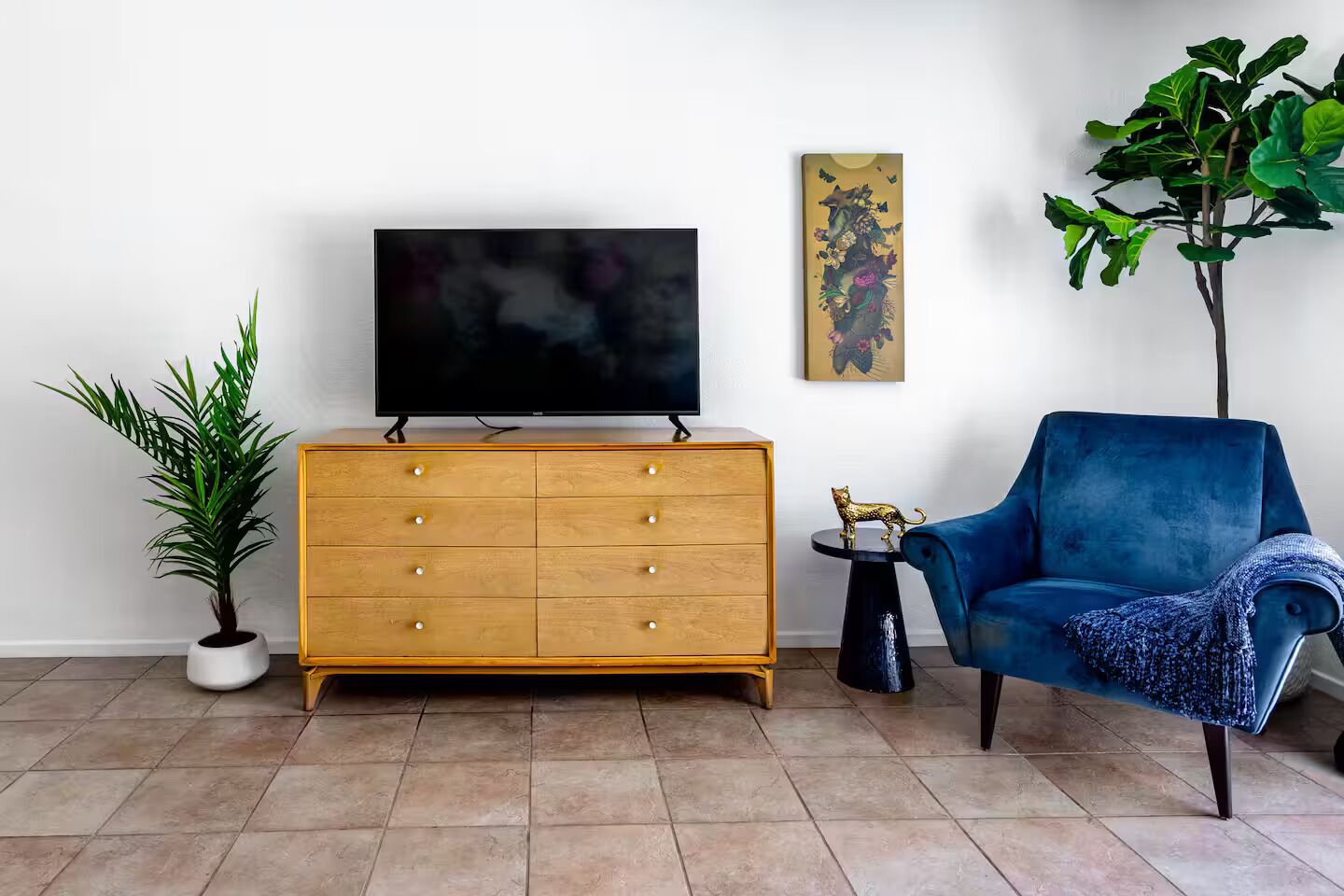 Een woonkamer met een houten kast, een blauwe fauteuil, een plant in pot, een televisie en een bijzettafeltje met een decoratief object.