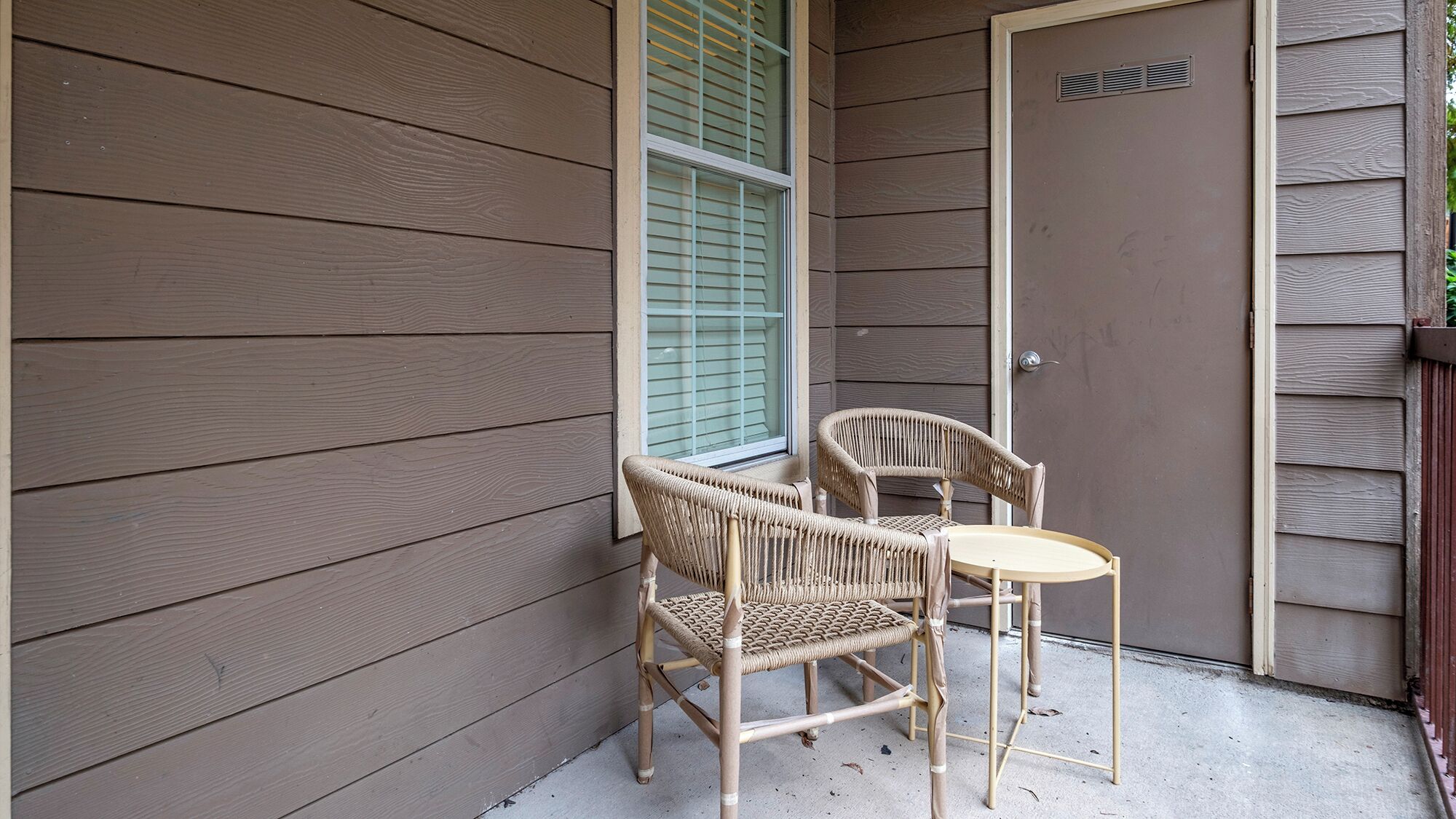A small outdoor seating area with two wicker chairs and a round table, positioned against a building with wooden siding.