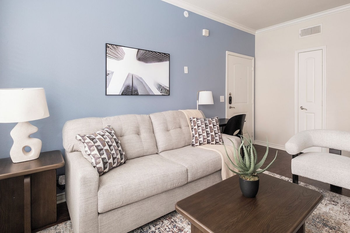 A living room with a beige sofa, patterned pillows, a wooden coffee table, a potted plant, and a framed picture on the wall.