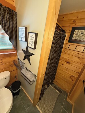 A bathroom with wooden paneling, a toilet, a sink, and a shower area. There are two framed signs on the wall, one with a star and the other with text. - Mountain Cabin Charm in Rumney with Wood Stove. (Rumney)