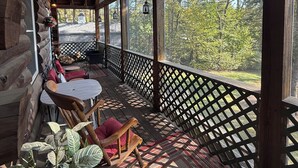 Una cabaña de troncos con porche cubierto, muebles de madera y vistas a árboles.