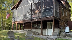 Una cabaña de troncos con un porche mosquitero, dos sillas Adirondack y un brasero.