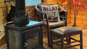Interior acogedor de una cabaña con estufa de leña, un sillón con cojín que dice 'RELAX' y una ventana con una bandera estadounidense.