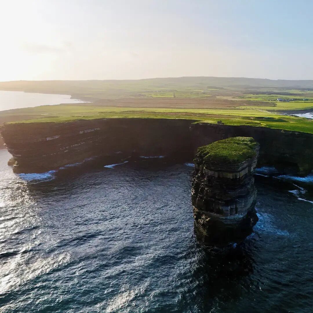 A coastal landscape with a prominent rock formation extending into the sea, surrounded by green fields and a distant shoreline.