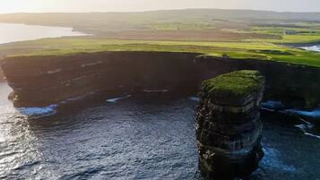 A coastal landscape with a prominent rock formation extending into the sea, surrounded by green fields and a distant shoreline.