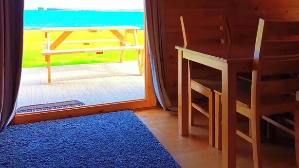A room with a wooden interior, a TV mounted on the wall, a bench outside the door, and a view of the beach. - Ceide Glamping  (Ballycastle)
