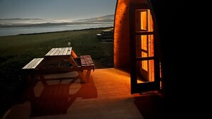 A cozy, semi-open structure with a wooden bench and table outside, a large open door, and a view of the sea under a bright moon. - Ceide Glamping  (Ballycastle)