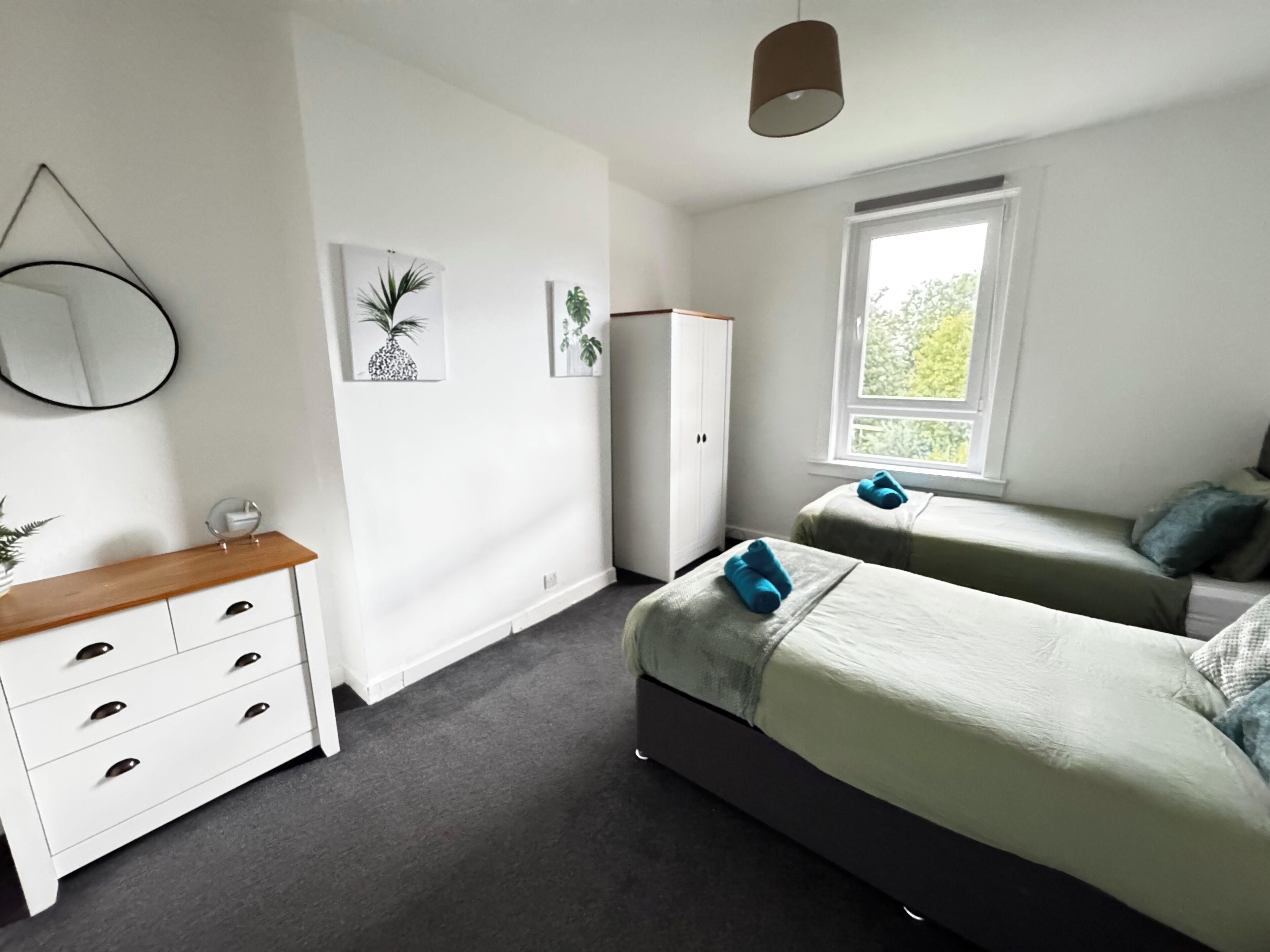 A bedroom with two beds, a wardrobe, a mirror, and a window with a view of greenery.