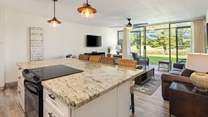 Une cuisine moderne avec un îlot central, un coin repas avec des chaises en osier, un téléviseur à écran plat et une grande porte-fenêtre coulissante donnant sur un jardin.