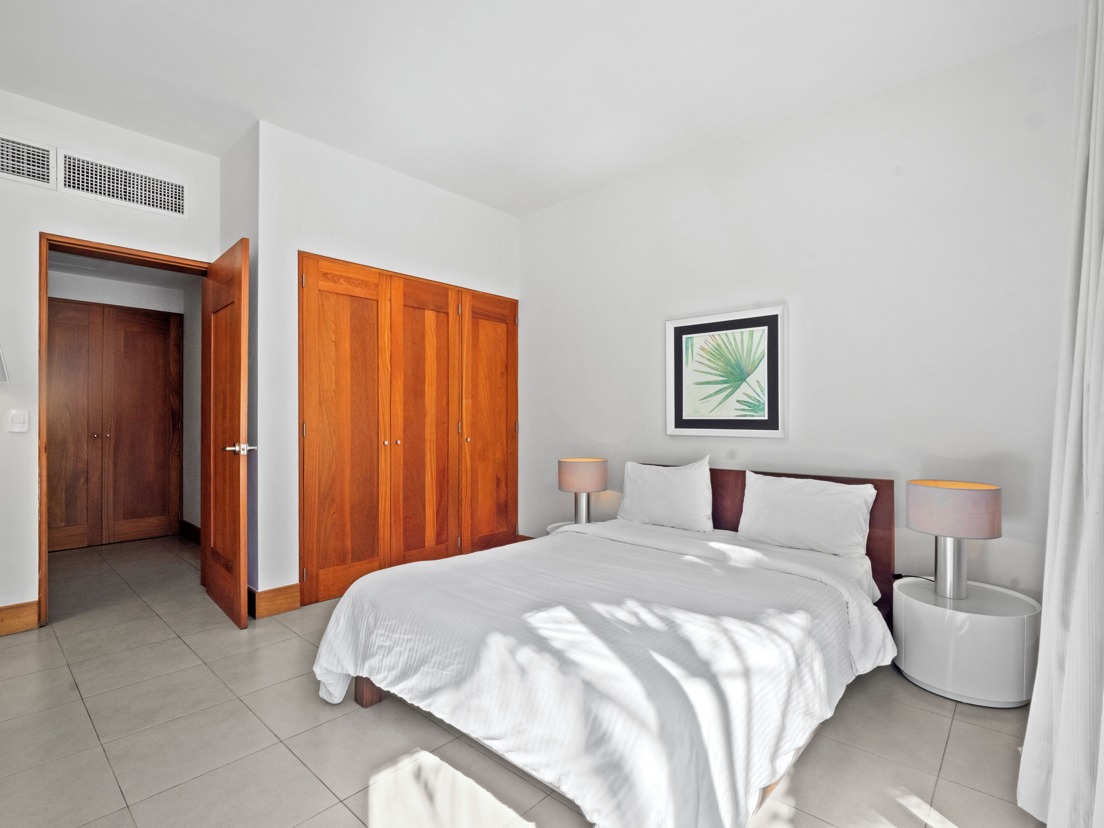 Quarto com cama, duas mesas de cabeceira, guarda-roupa de madeira, quadro emoldurado na parede e porta que dá para outro cômodo.