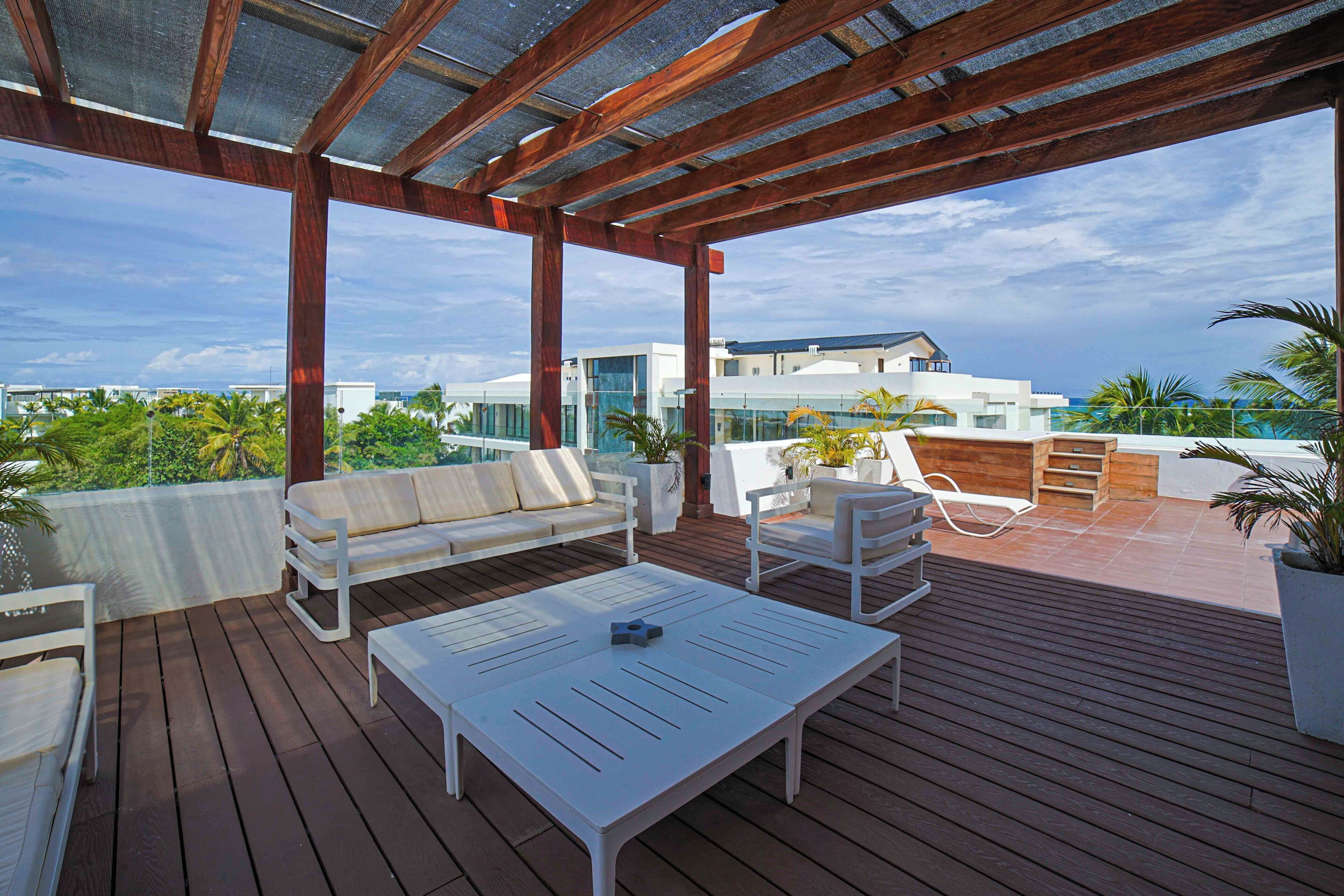 Un espacio exterior techado con un piso de madera, mobiliario blanco y vistas a edificios modernos y palmeras.