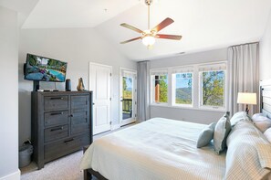 A bedroom with a bed, a dresser, a ceiling fan, a TV, and windows with a view of trees and hills. - Wintergreens R331- Mtn Condo/View, Gated Resort, Indoor/Outdoor Pools & Hot Tub (Boone)