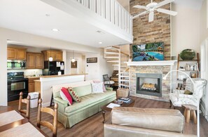 A spacious living area with a fireplace, a high ceiling with a fan, and a kitchen in the background. - Echota Wintergreens 1632- Mtn Condo, Gated Resort, Indoor/Outdoor Pools & Hot Tu (Boone)