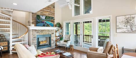 A spacious living room with a fireplace, a staircase, and a balcony with a view of greenery.