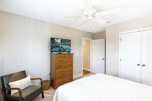 Une chambre à coucher avec un lit, une commode en bois, un téléviseur posé dessus, un ventilateur de plafond et une porte.