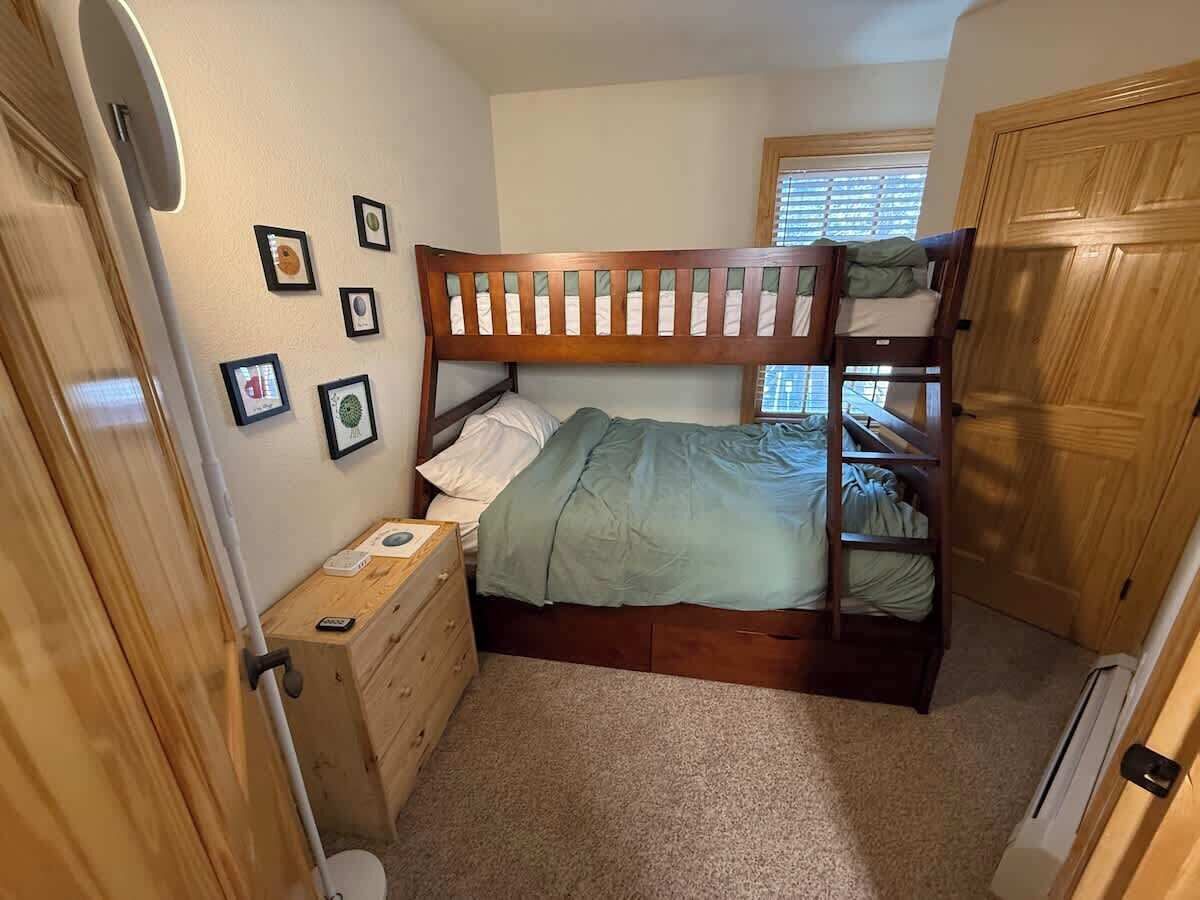 A small, cozy bedroom with a bunk bed, a wooden dresser, and a window with blinds.