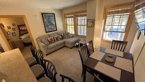 A living room with a sofa, a dining table with chairs, and a large window with blinds.