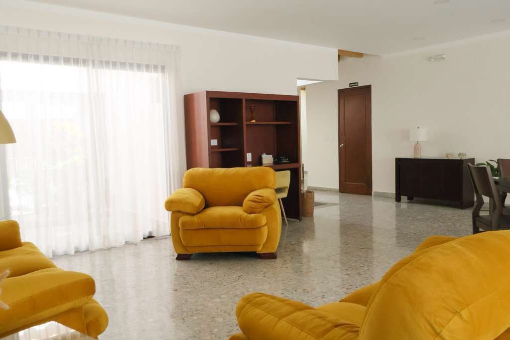 Una sala de estar con un sillón amarillo, una estantería de madera y un comedor con una mesa y sillas.