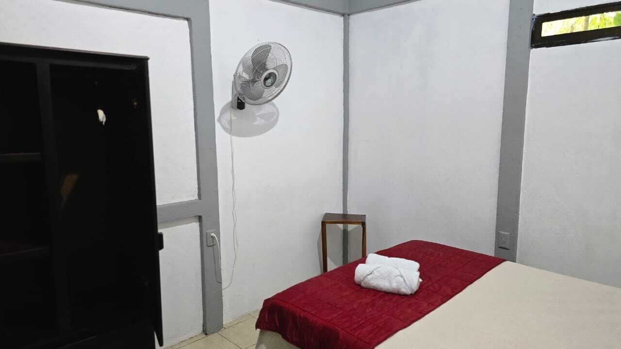 Una habitación pequeña con una cama individual, una colcha roja, un ventilador de pie y una puerta.
