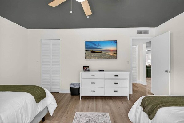Une chambre avec deux lits, une commode, un ventilateur de plafond, un tableau et une porte donnant sur une autre pièce.