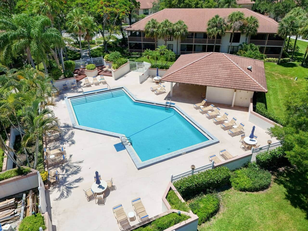 A swimming pool with lounge chairs and a covered area, surrounded by greenery and a building with a tiled roof.