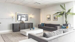 A modern living room with a grey sofa, a white coffee table, and a grey cabinet. There are two wall-mounted lights and a large plant. - private balcony ON THE BEACH with pool 24hr front desk parking spot laundry gym (Hallandale Beach)